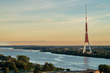 Sunset view of the television tower located on the banks of the Daugava River, with warm orange hues filling the sky. The scene captures a serene and atmospheric moment.