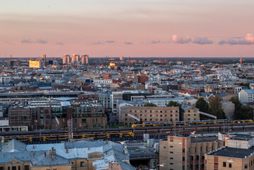Naklejka premium Panoramic cityscape featuring a railway station in the foreground, with train tracks, platforms, and multiple trains leading toward the urban skyline in the distance.