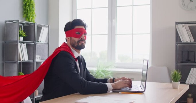 Focused businessman working with laptop in office, wearing superhero mask and costume, showcasing business power and strength, enhancing a unique atmosphere in a professional space of dedication.
