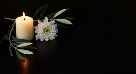 A lit candle accompanied by a white flower and green leaves on a dark surface.