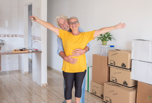 Cheerful senior couple moving into a new home, surrounded by moving boxes in an empty room. The woman has outstretched arms holding house keys, symbolizing their new beginning as retirees