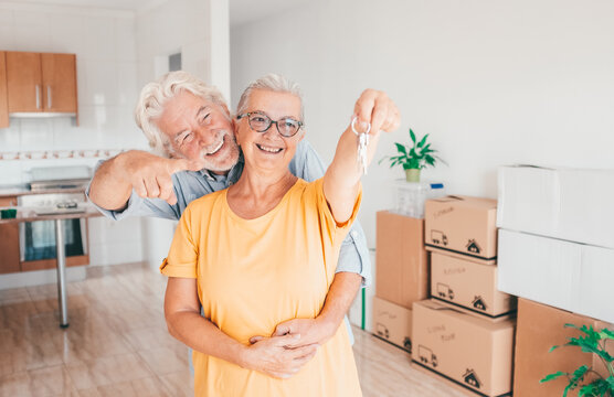 Cheerful senior couple hug moving into a new home, surrounded by moving boxes in an empty room. The woman holding house keys, excited for their new beginning, real estate, property concept