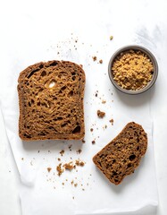 Two slices of dark bread, crumbs, and a small bowl of coarse crumbs on parchment paper