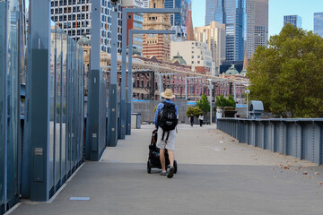 A father pushes a stroller across a pedestrian bridge with Melbourne CBD skyline as the background, capturing urban family life.