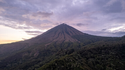 Fototapeta premium A breathtaking view of Mount Inerie in Flores, Indonesia, during sunset. The volcanic peak rises steeply with clouds at the summit, surrounded by lush green forest and dramatic sky. 