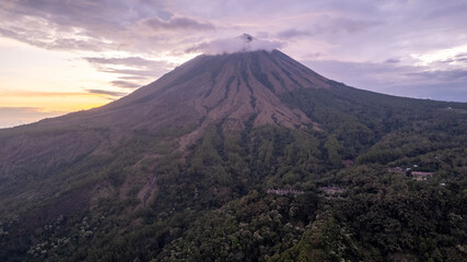 Fototapeta premium A breathtaking view of Mount Inerie in Flores, Indonesia, during sunset. The volcanic peak rises steeply with clouds at the summit, surrounded by lush green forest and dramatic sky. 