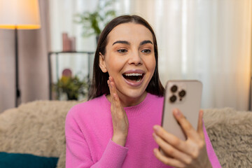 Young woman at home looking at mobile phone screen opening mouth in surprise reacting emotionally....