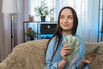 Young woman at home happily sitting on sofa counting large pile stack of dollar cash bills, celebrating wealth. Caucasian girl joyful managing successful personal budget, satisfied financial profit