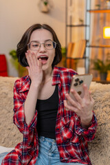 Young woman looks at smartphone on home sofa, reacting with joy after receiving good news and lottery win. Girl celebrates online jackpot with victory gesture and visible happiness and satisfaction