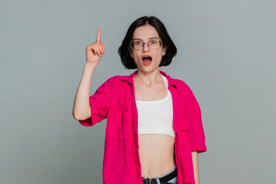 Young woman touches chin while thinking, then smiles and lifts finger up with excitement after finding idea or solution. Caucasian girl shows realization and inspiration, isolated on gray background.