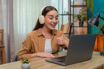 Young woman sitting at home table wears wireless headphones and speaks on laptop video call giving support. Caucasian girl keeps serious tone gestures calmly giving clear helpful answers to customers