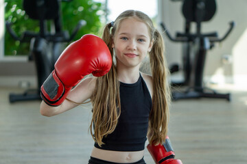 A young girl is posing with oversized red boxing gloves in a well-lit gym. She exudes confidence...