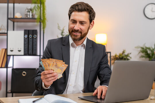 Middle-aged businessman at home office counts cash euros and enters amounts on laptop for daily budget well. Freelancer guy at table writes notes in notebook adds sums and plans expenses and savings.
