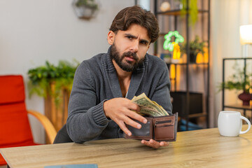 Indian man at home opens wallet finds only coins looks sad thinks about debt and overdue bills low pay. Arabian guy at desk plans budget counts money notes small income reads fees plans reduce spend