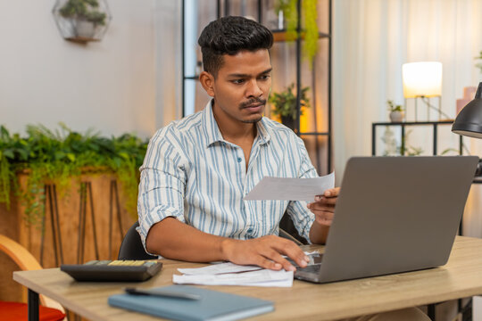 Indian man at home uses laptop and calculator enters bills tracks expenses sets budget and savings. Arabian guy at desk sorts receipts checks invoice and tax uses spreadsheet plans budget save money