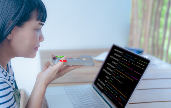 Asian woman using voice command on smartphone while working on laptop with code on screen. Concept of multitasking, voice technology, remote work, AI assistant, and programming