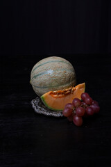 Top view of green cantaloupe melon on a tray, green leaves and red grapes, black background, vertical, with copy space