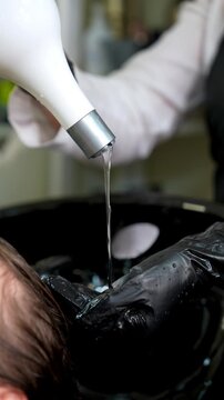 Professional hairstylist washing clients hair in salon sink. Black gloves, shampoo from white bottle, modern hair care process, beauty treatment. 