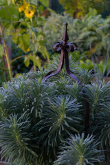 A decorative metal garden obelisk finial sits nestled among spiky, blue-green foliage with a yellow sunflower blurred in the background on a sunny day.