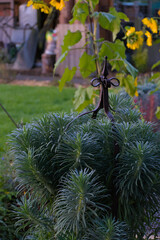 A decorative metal garden obelisk finial sits nestled among spiky, blue-green foliage with a yellow sunflower blurred in the background on a sunny day.