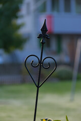 A black wrought iron garden stake with a decorative heart and a fleur-de-lis top stands in a grassy yard with a blurred, misty background.