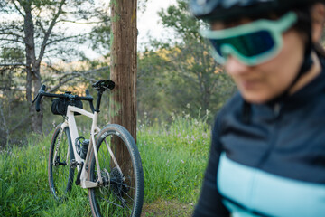 Young woman cyclist resting near gravel bike leaning against wooden pole in nature