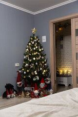 Christmas tree decorated with ornaments and lights in cozy home interior with gray walls, gifts, and Scandinavian gnomes near the doorway