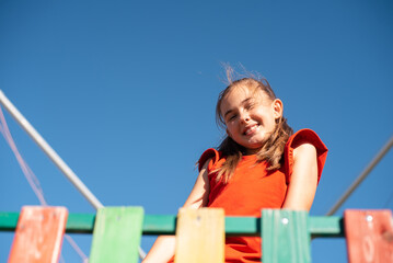 A bright, smiling child girl plays on a colorful wooden playground against a clear blue sky, enjoying a fun and active outdoor moment