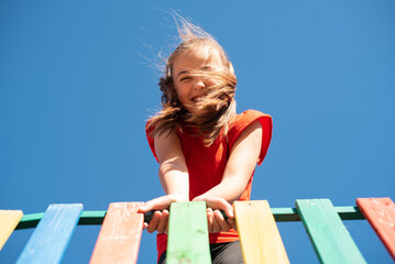 A bright, smiling child girl in a red shirt plays on a colorful wooden playground against a clear blue sky, with her windy hair covering her face, enjoying a fun and active outdoor moment