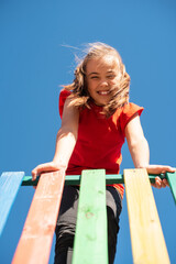 Bright smiling child in red shirt playing on colorful wooden playground against clear blue sky, joyful summer outdoor activity.