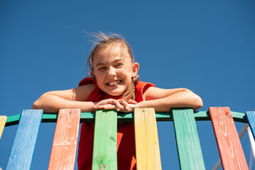 A smiling child in red shirt playing on colorful wooden playground against clear blue sky, enjoying a fun and active outdoor moment.