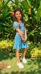 An 8-year-old girl in a tropical outfit stands smiling among lush green plants in Bali, with a red tropical flow Bright natural light, vibrant colors, and a cheerful warm atmosphere. Summer activities