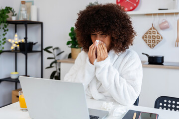 Sick young multiracial woman working on laptop in the kitchen at home, blowing nose and sipping juice at kitchen table. Dedicated freelancer despite illness