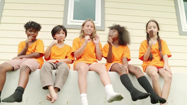 Group young male and female international school students sit in a row eating popsicles to cool down and enjoy their favorite delicious cool ice cream after playing outdoor sports during the summer.