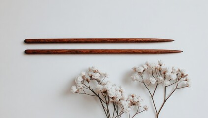 Two parallel wooden knitting needles rest against a backdrop of delicate white flowers, showcasing a simple yet elegant craftsmanship.