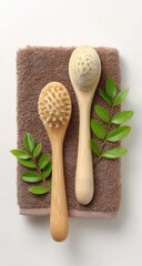 Two natural bristle body brushes with light brown wooden handles sit atop a taupe towel, accented by fresh green leaves.