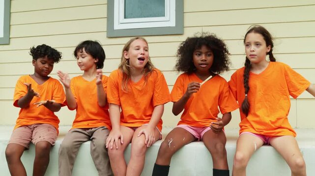Group young male and female international school students sit in a row eating popsicles to cool down and enjoy their favorite delicious cool ice cream after playing outdoor sports during the summer.