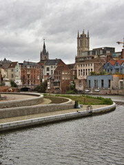 Obraz premium Riverbend with Historic Towers in Ghent - A bend in the Leie River with a view of traditional houses and two historic church spires under a cloudy sky.