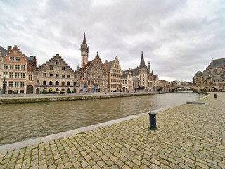 The scenic waterfront of Ghent features the historic guild houses and buildings lining the Lys river under a dramatic, cloudy sky, with a cobblestone street in the foreground.