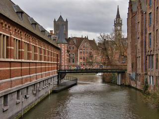  A unique perspective of a canal in Ghent, framed by two rows of historical buildings, a small pedestrian bridge, and the clock tower of the old Post Office in the distance.