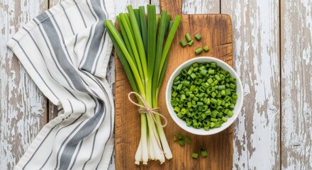 Overhead view of fresh green onions tied on a wooden board with chopped onions in a bowl and striped towel