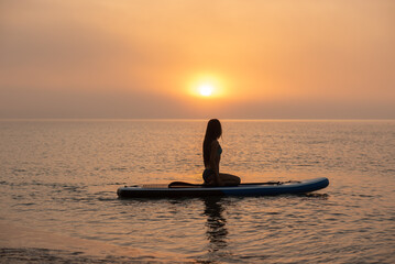 A beautiful, slender, athletic girl is relaxing on a SUP in the open sea at dawn on a sunny summer day.