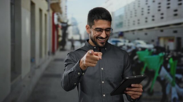 Smiling young man using a tablet and pointing confidently on an urban street, exuding charm and friendliness in an outdoor city environment.