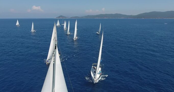 Aerial view of a sailing regatta with multiple yachts competing on a sunny day in the open sea, surrounded by scenic coastlines