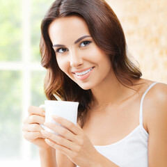 Portrait of happy smiling brunette cute woman in white lingerie top holding mug cup, drinking coffee or tea, at home, near window. Square image