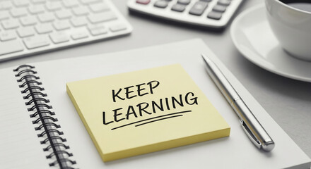 Yellow sticky note with handwritten text "Keep Learning" on a notepad, near keyboard, calculator, pen, and coffee cup, suggesting continuous education and self-improvement