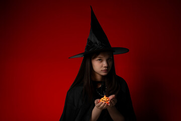smilling creepy girl wearing a witch hat and black cape presents a small jack-o'-lantern against a...