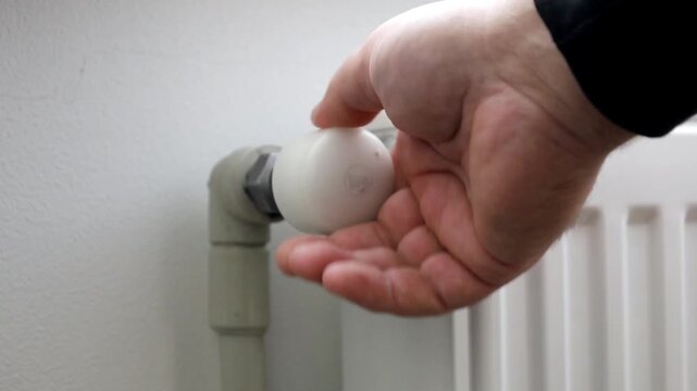 A close-up shot of a person turning the valve on a shut-off tap of a white heating radiator. This process of temperature regulation is a key element of a modern indoor heating system, ensuring comfort