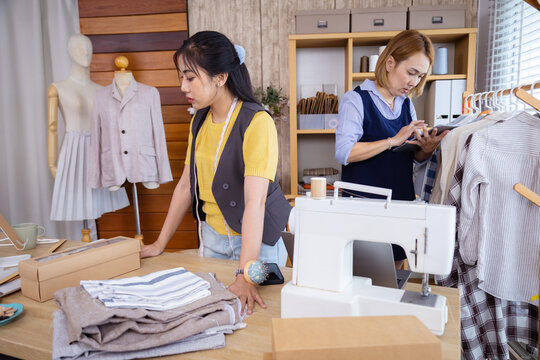 Two female partners running clothing shop together, one using phone and laptop to receive order while other checking garments, teamwork showing cooperation in small business startup fashion store