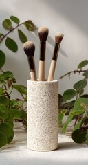 Three makeup brushes, displayed in a speckled, light beige cylinder vase, sit against a bright backdrop, evoking a sense of simple elegance.